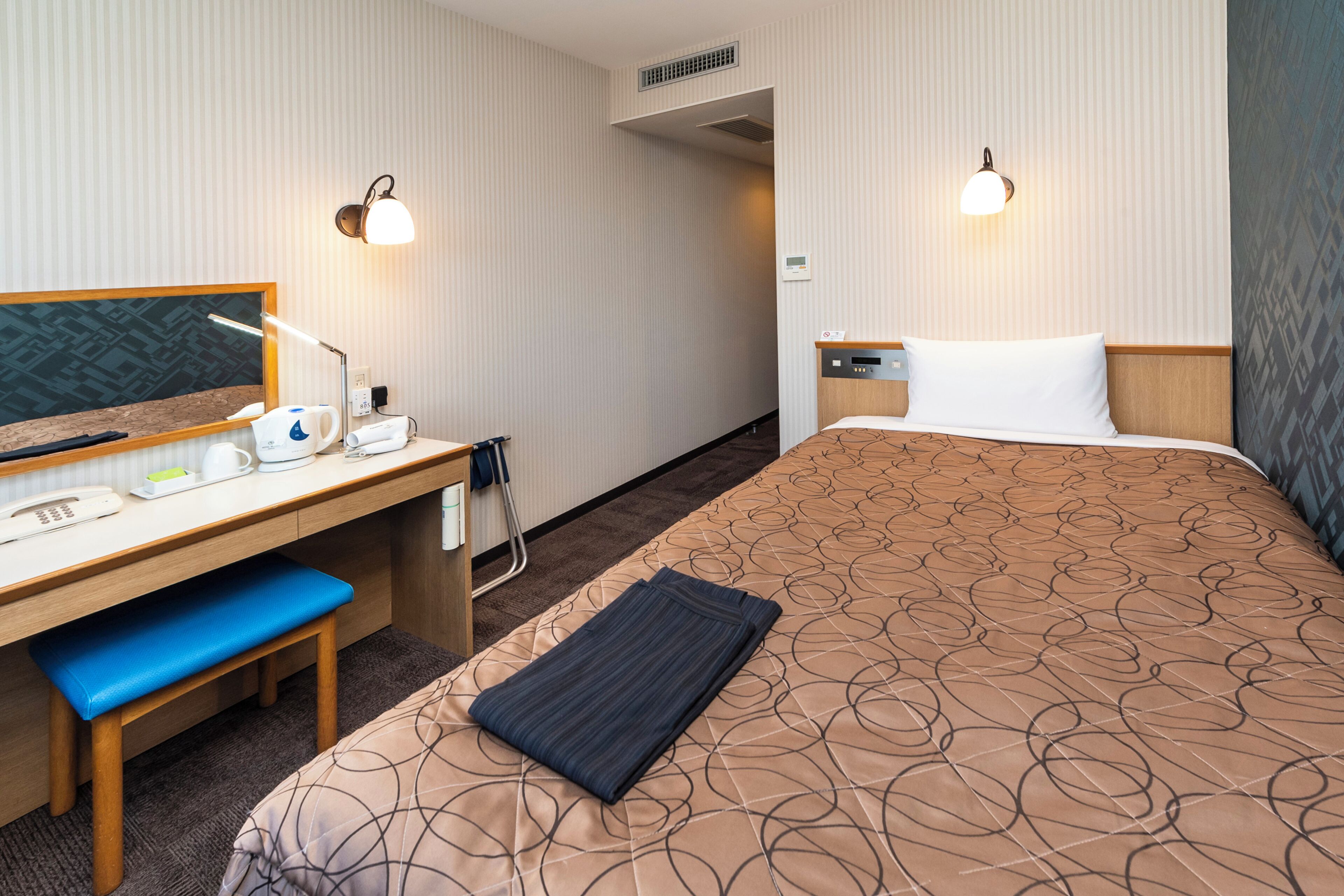 A hotel room with a bed, desk, chair, and two wall-mounted lamps.