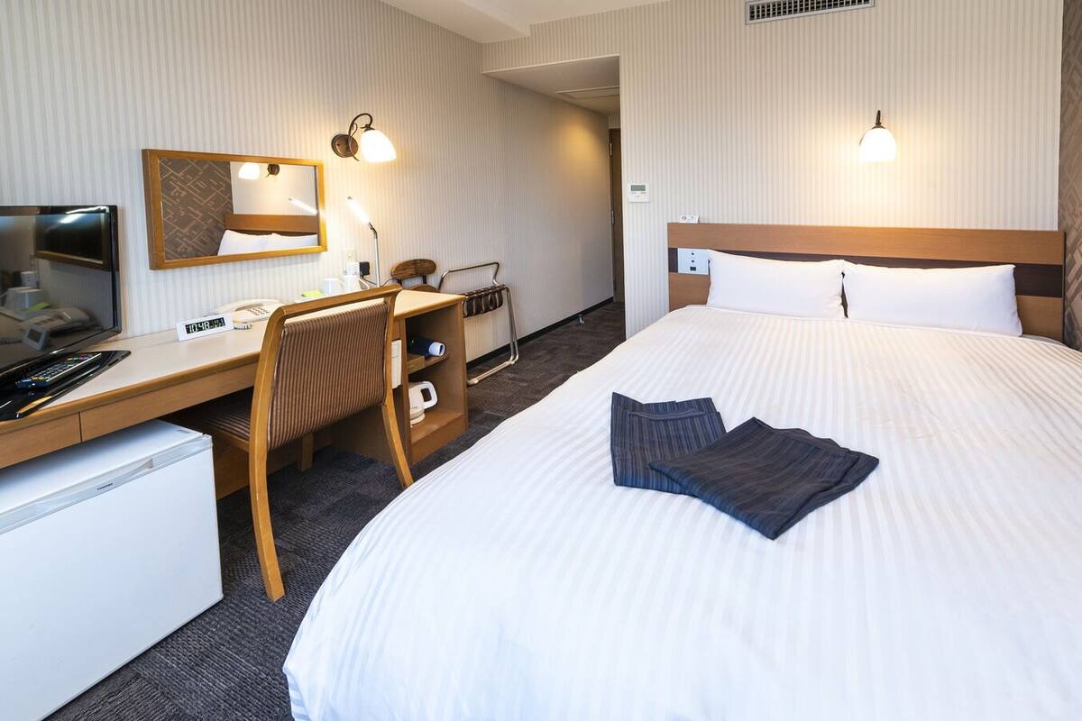 A hotel room with a bed, desk, chair, and a small refrigerator.