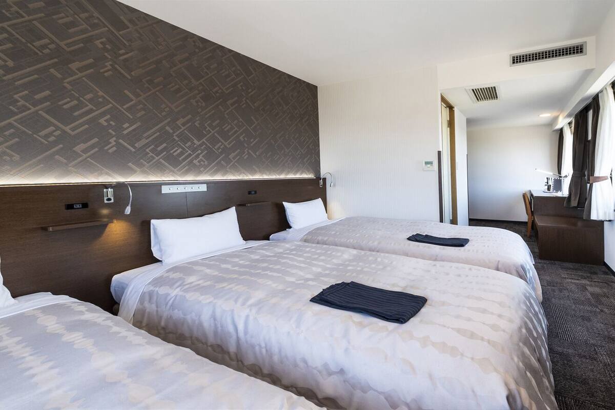 A hotel room with three beds, a wooden headboard, a desk, and a chair.