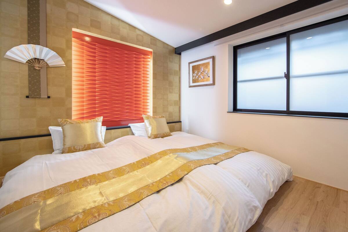 Ein Schlafzimmer mit einem großen Bett, einem Fenster mit Jalousien, einem Wandventilator und einem gerahmten Bild.