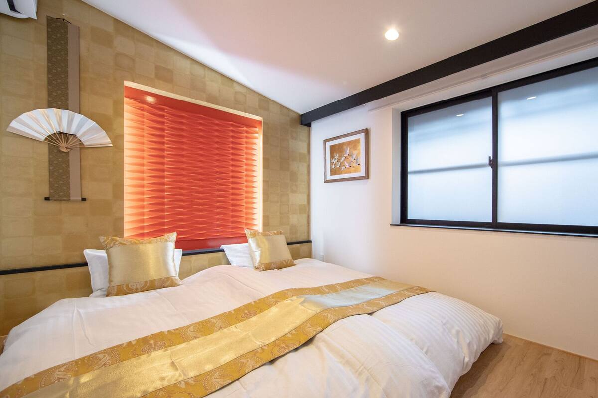 Ein Schlafzimmer mit einem großen Bett, einem Fenster mit Jalousien, einem Wandventilator und einem gerahmten Bild.