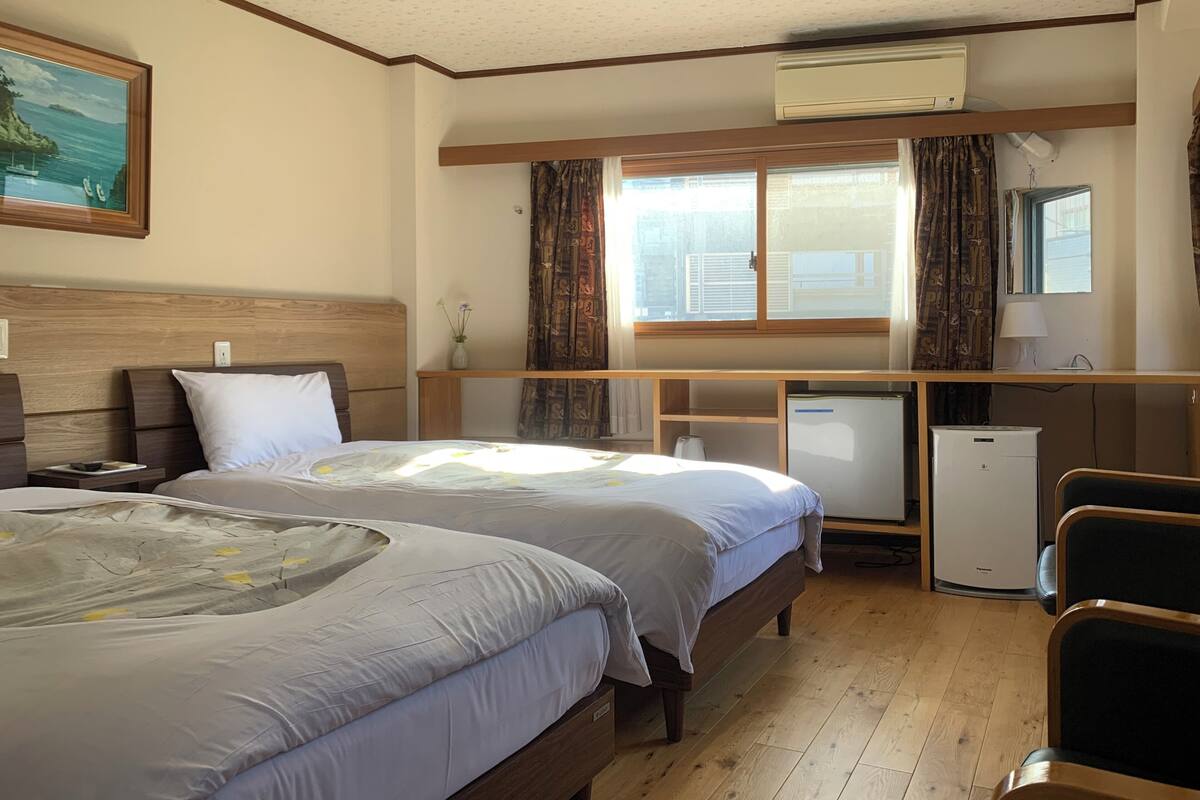 A hotel room with two beds, a desk, a refrigerator, and a window with curtains.