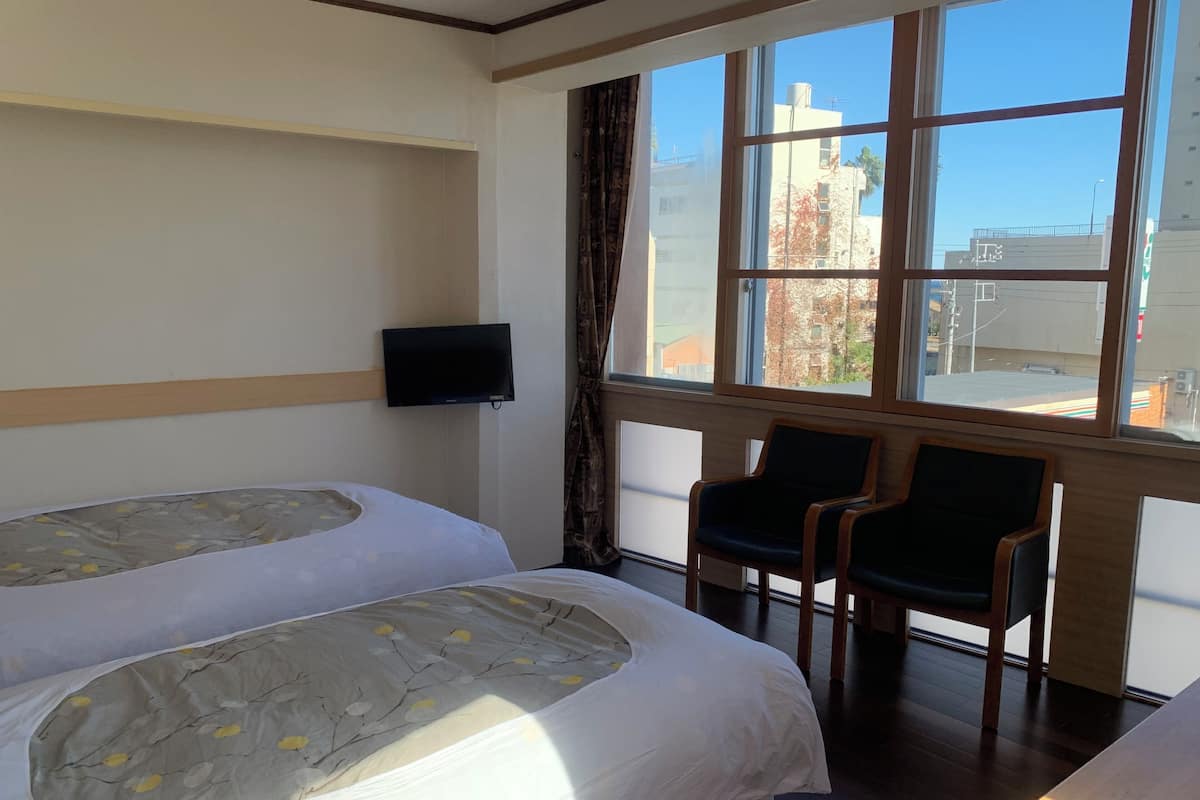 A hotel room with two beds, a TV, and a window with a view of buildings.