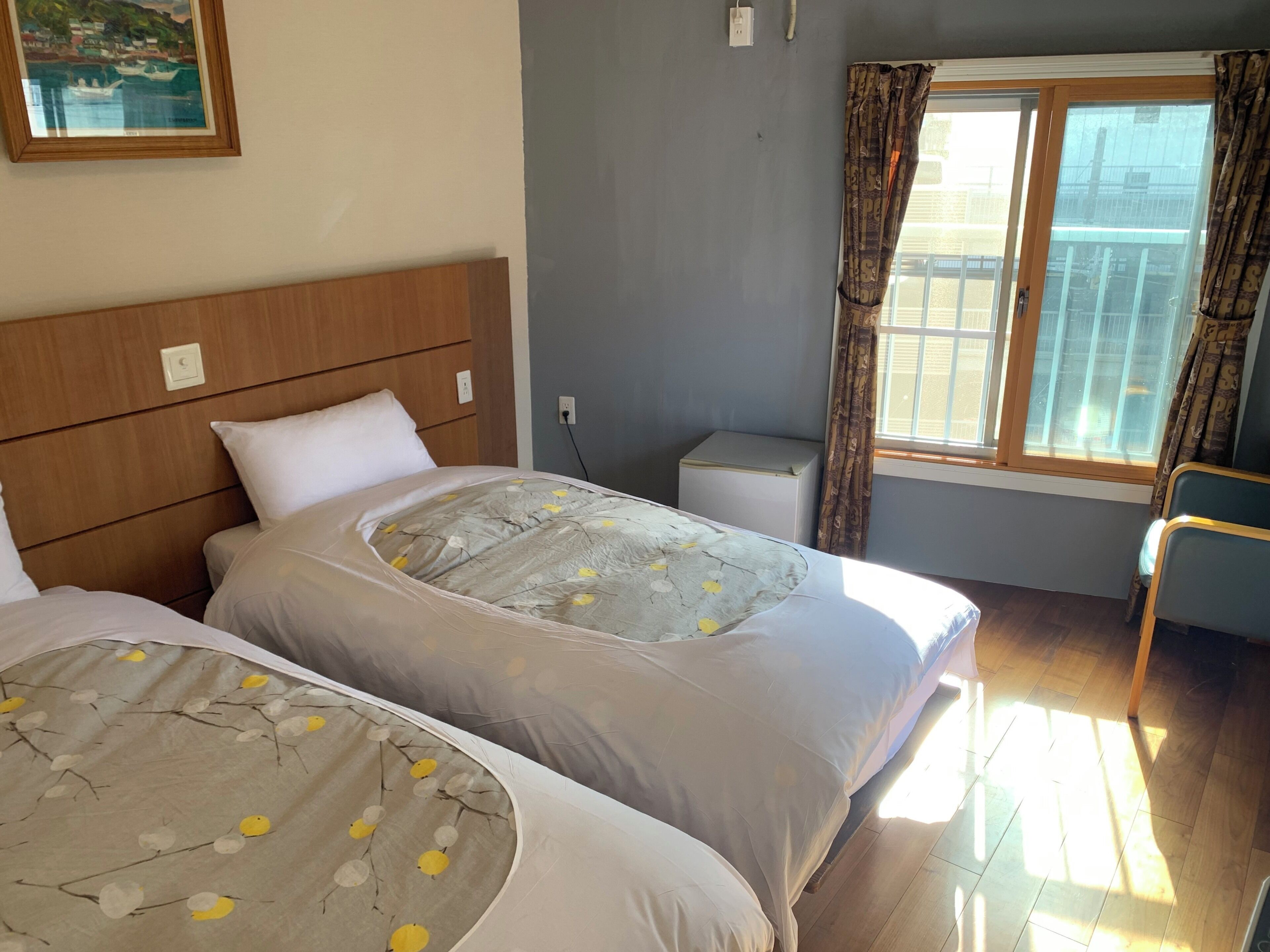 A hotel room with two beds, a small bedside table, a window with curtains, and a painting on the wall.