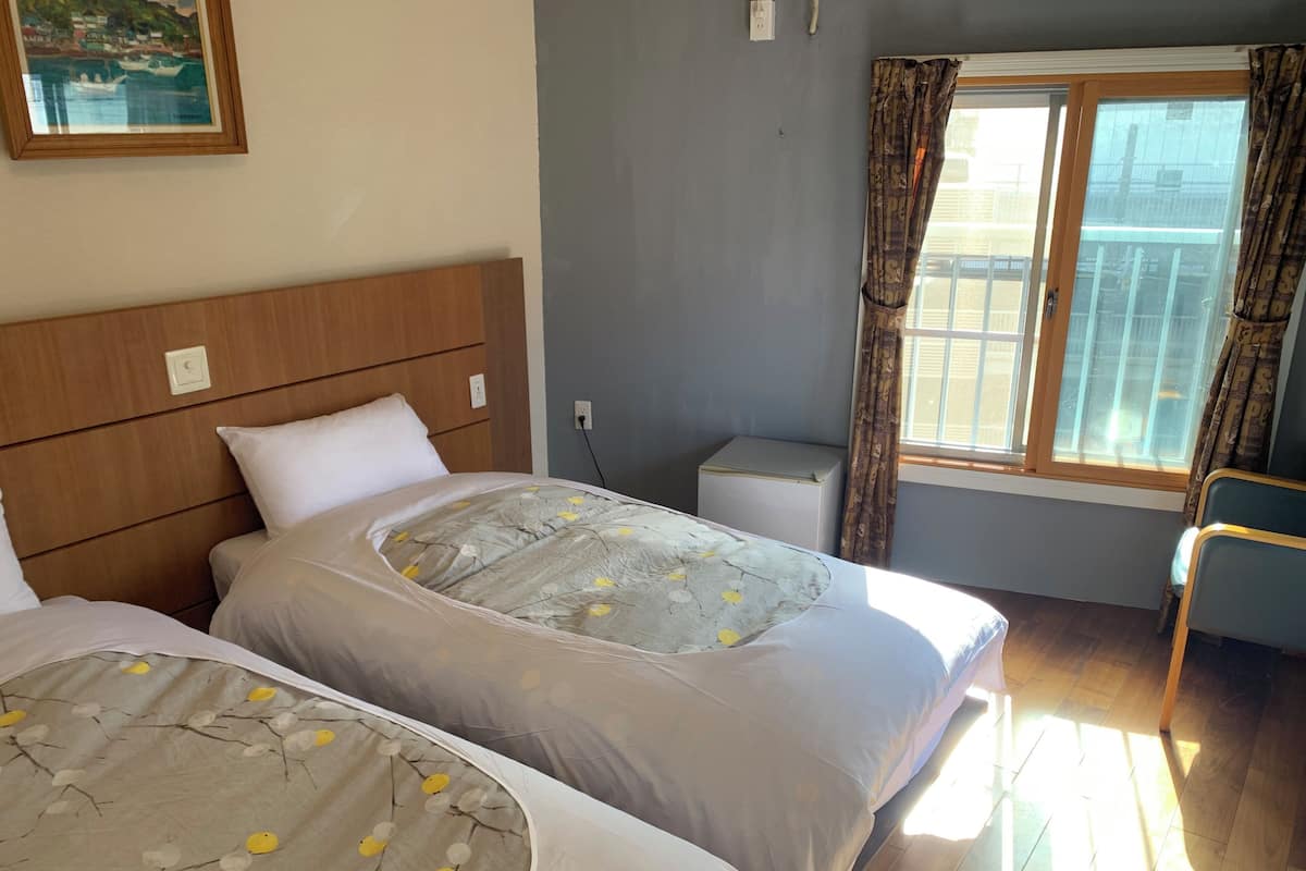 A hotel room with two beds, a small bedside table, a window with curtains, and a painting on the wall.