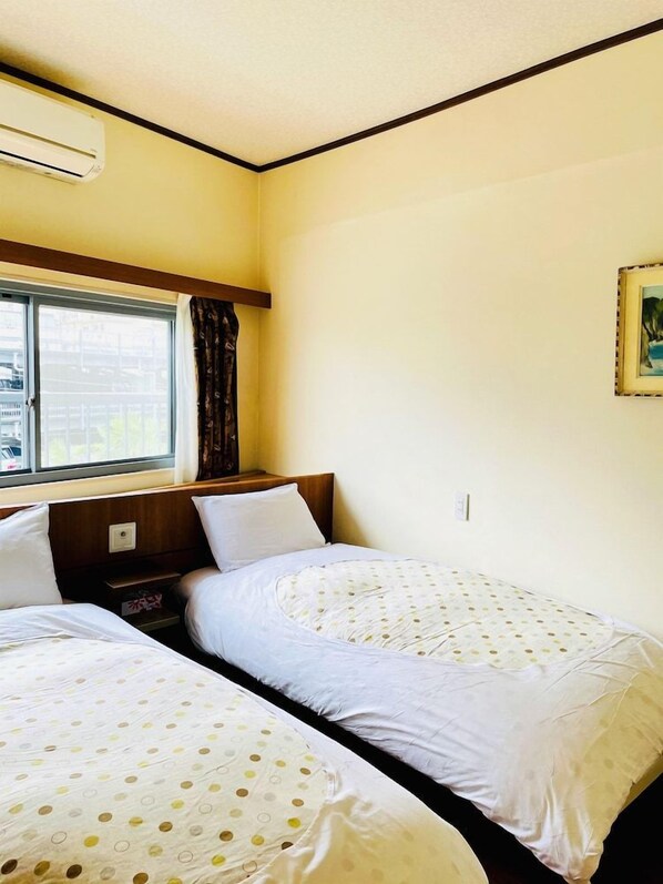 A room with two beds, a window with curtains, an air conditioning unit, and a framed picture on the wall.
