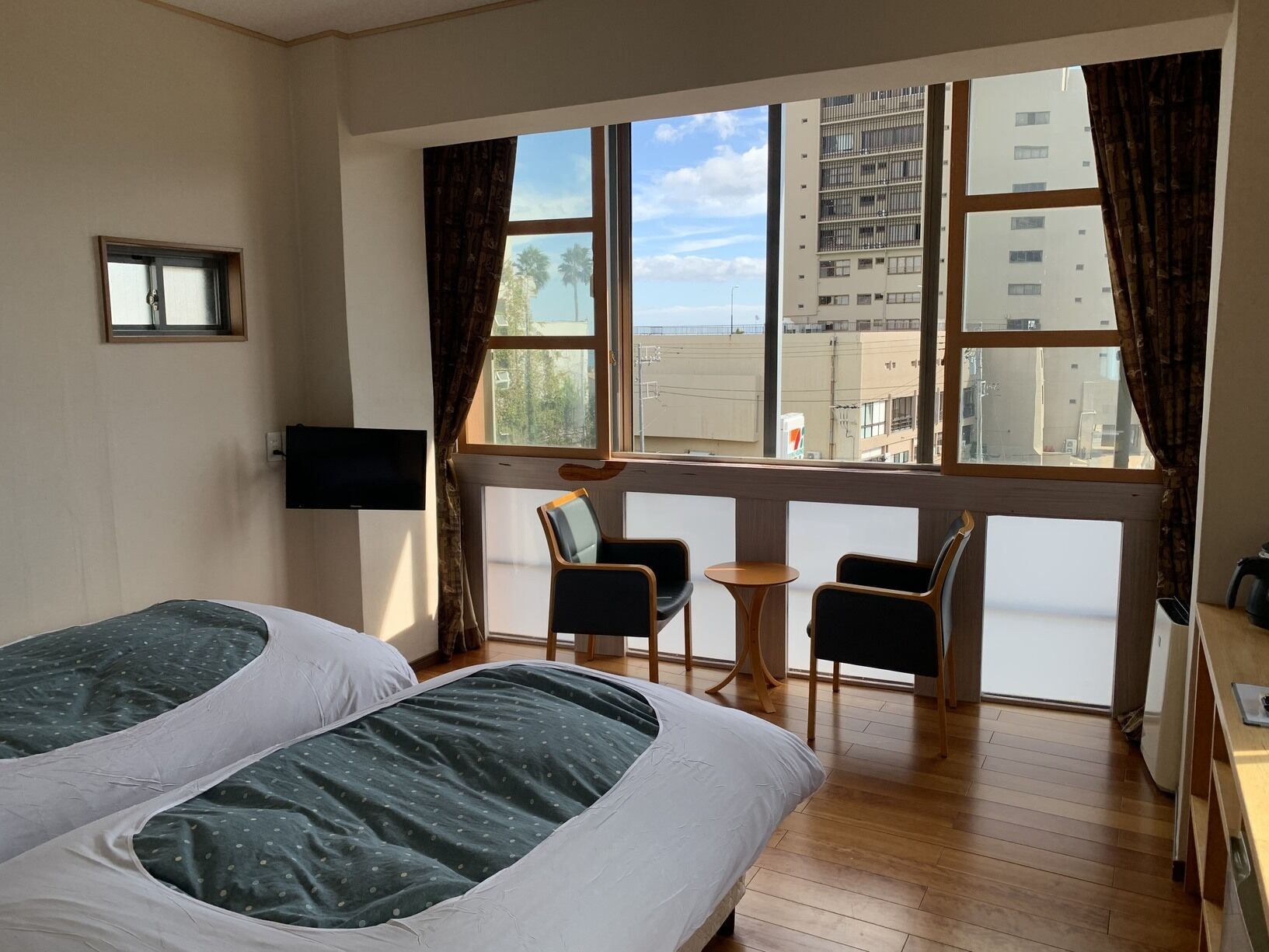 A hotel room with two beds, a TV, a small table, two chairs, and a large window with a view of buildings.