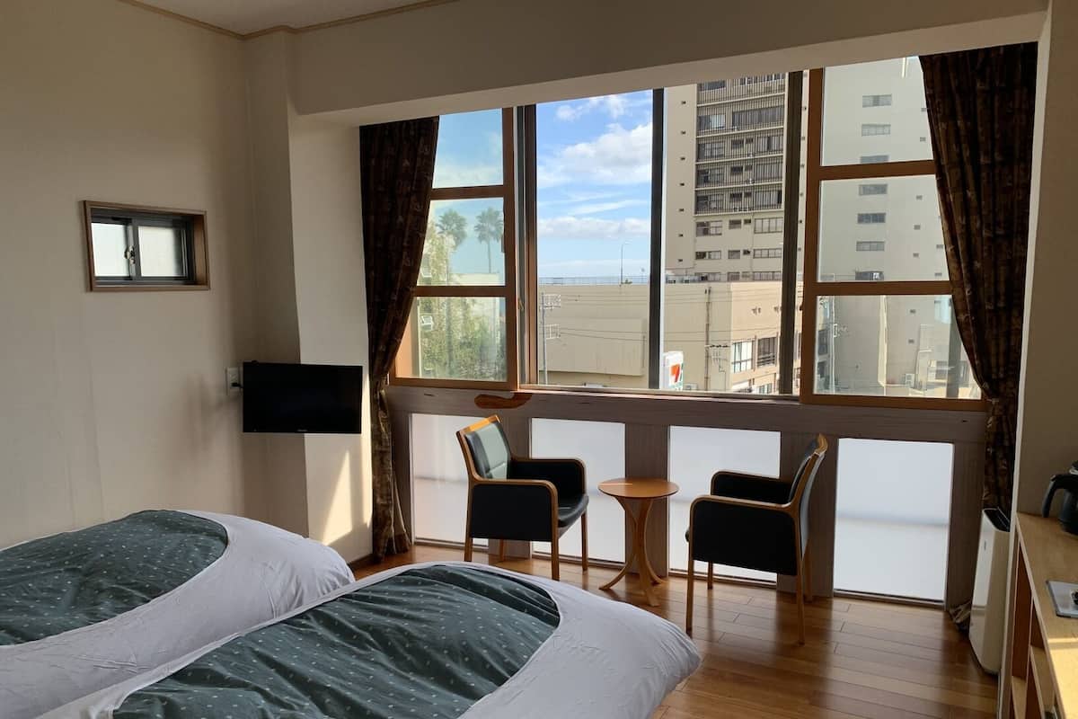 A hotel room with two beds, a TV, a small table, two chairs, and a large window with a view of buildings.