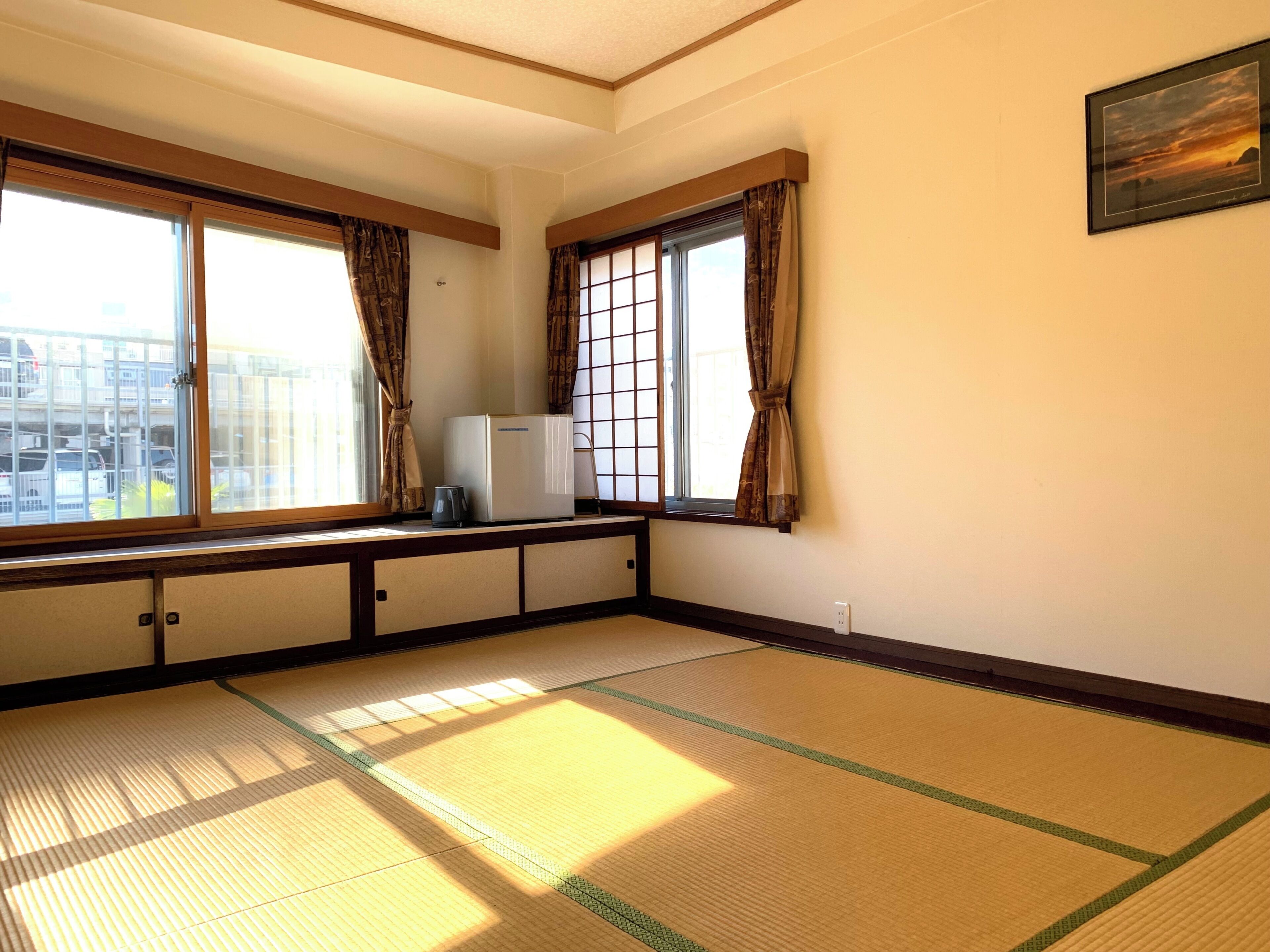 A room with tatami flooring, large windows with curtains, a small refrigerator, and a framed picture on the wall.