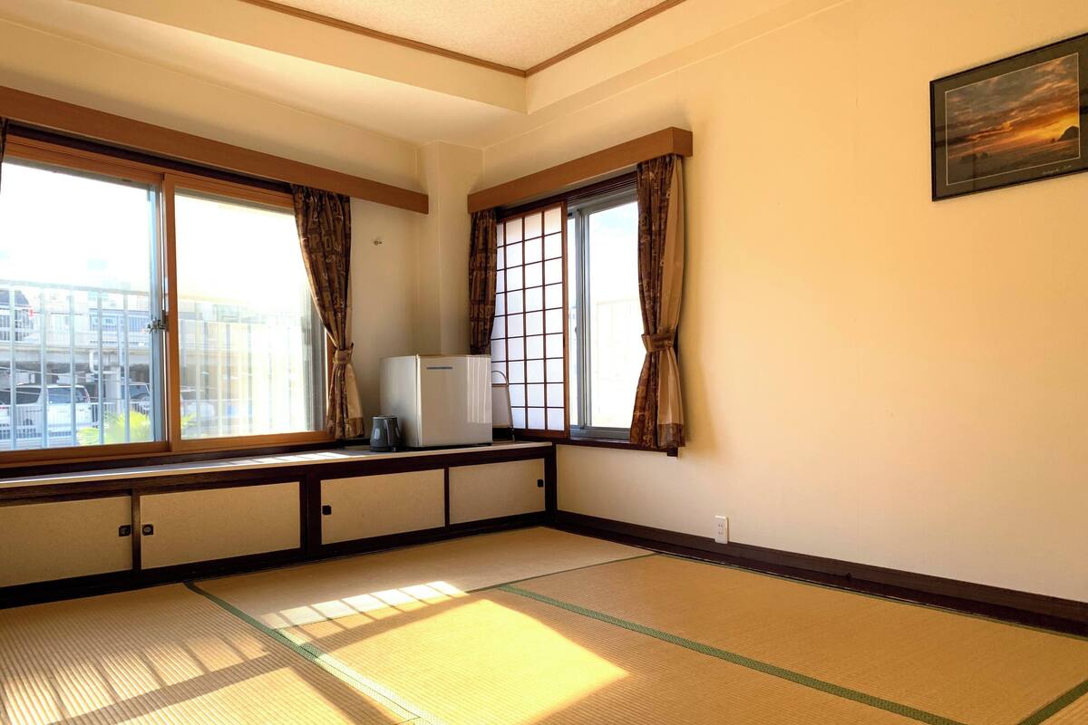 A room with tatami flooring, large windows with curtains, a small refrigerator, and a framed picture on the wall.