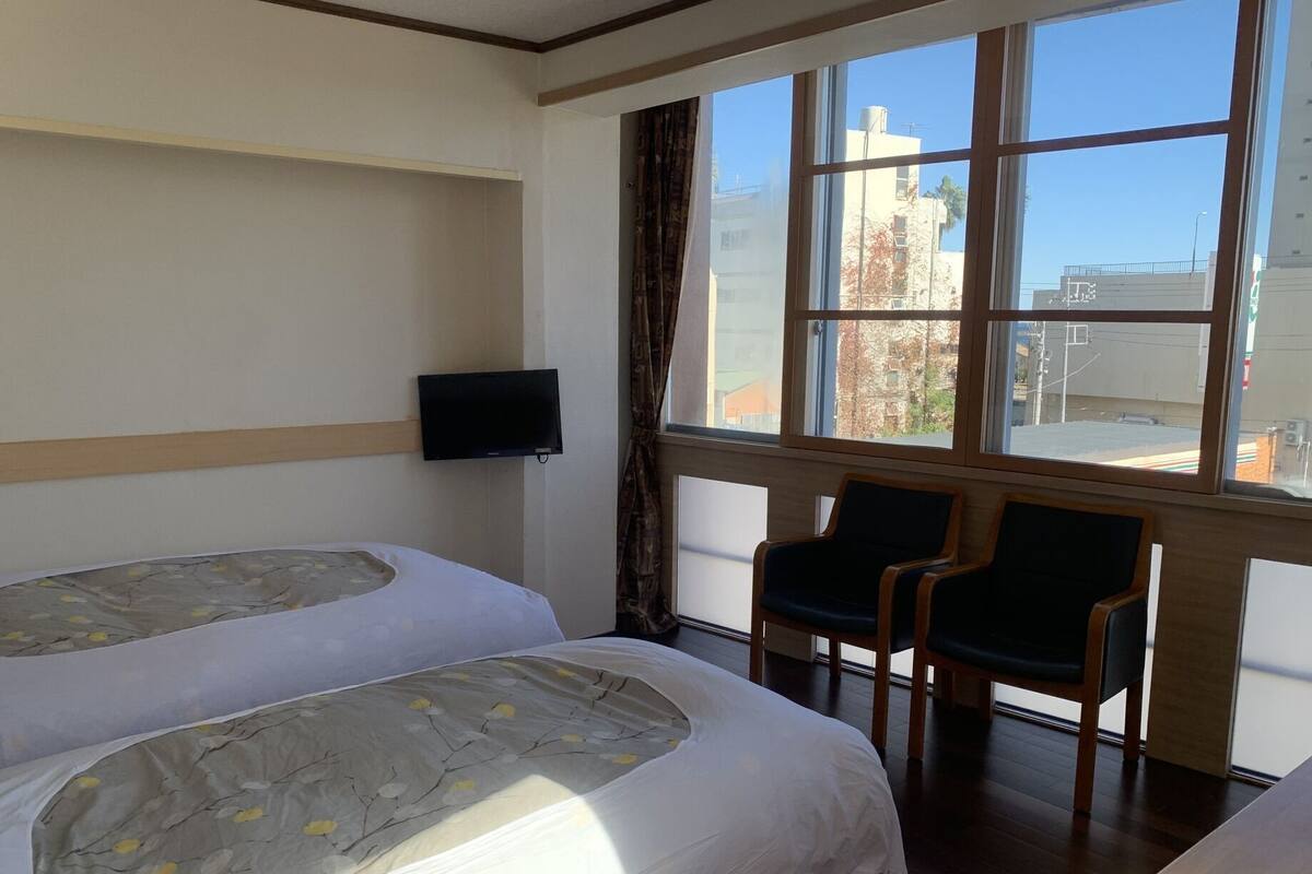A hotel room with two beds, a TV, a window with a city view, and two chairs.