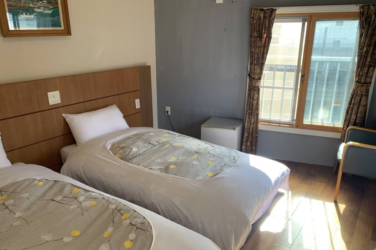 A hotel room with two beds, a small bedside table, a window with curtains, and a painting on the wall.