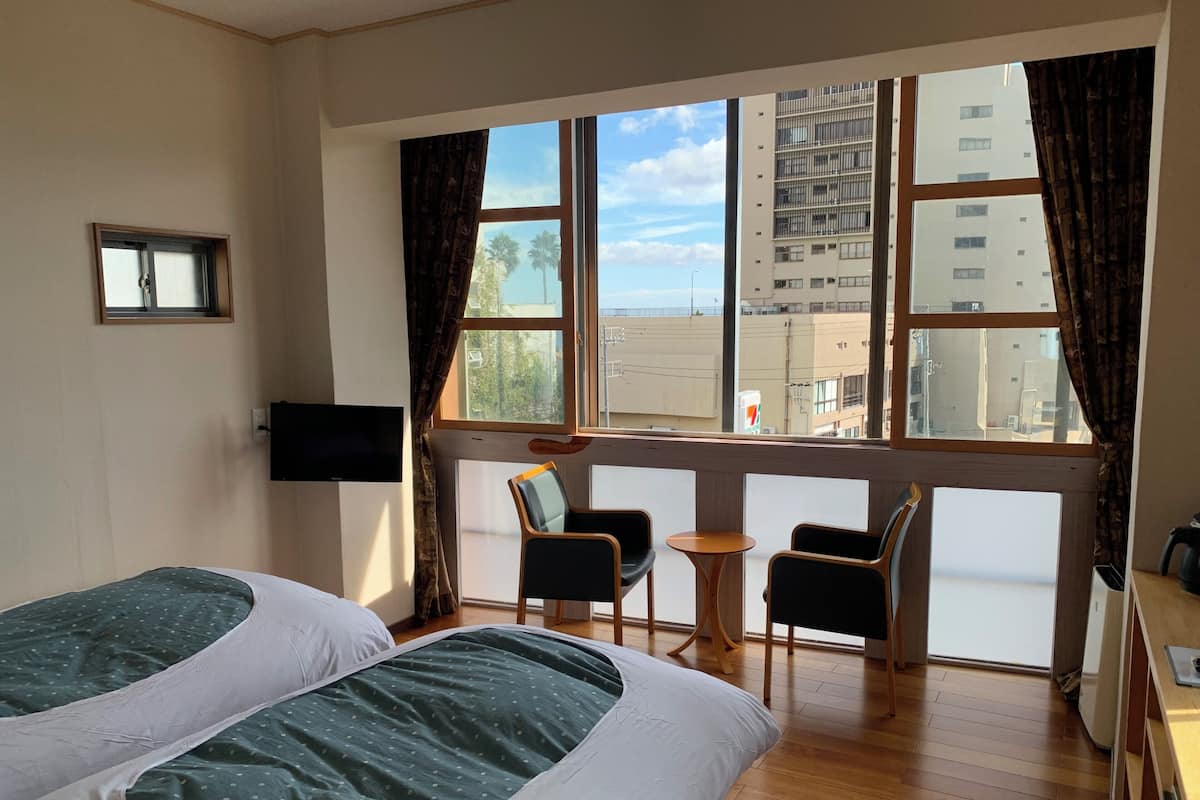 A hotel room with two beds, a TV, a small table, two chairs, and a large window with a view of buildings.