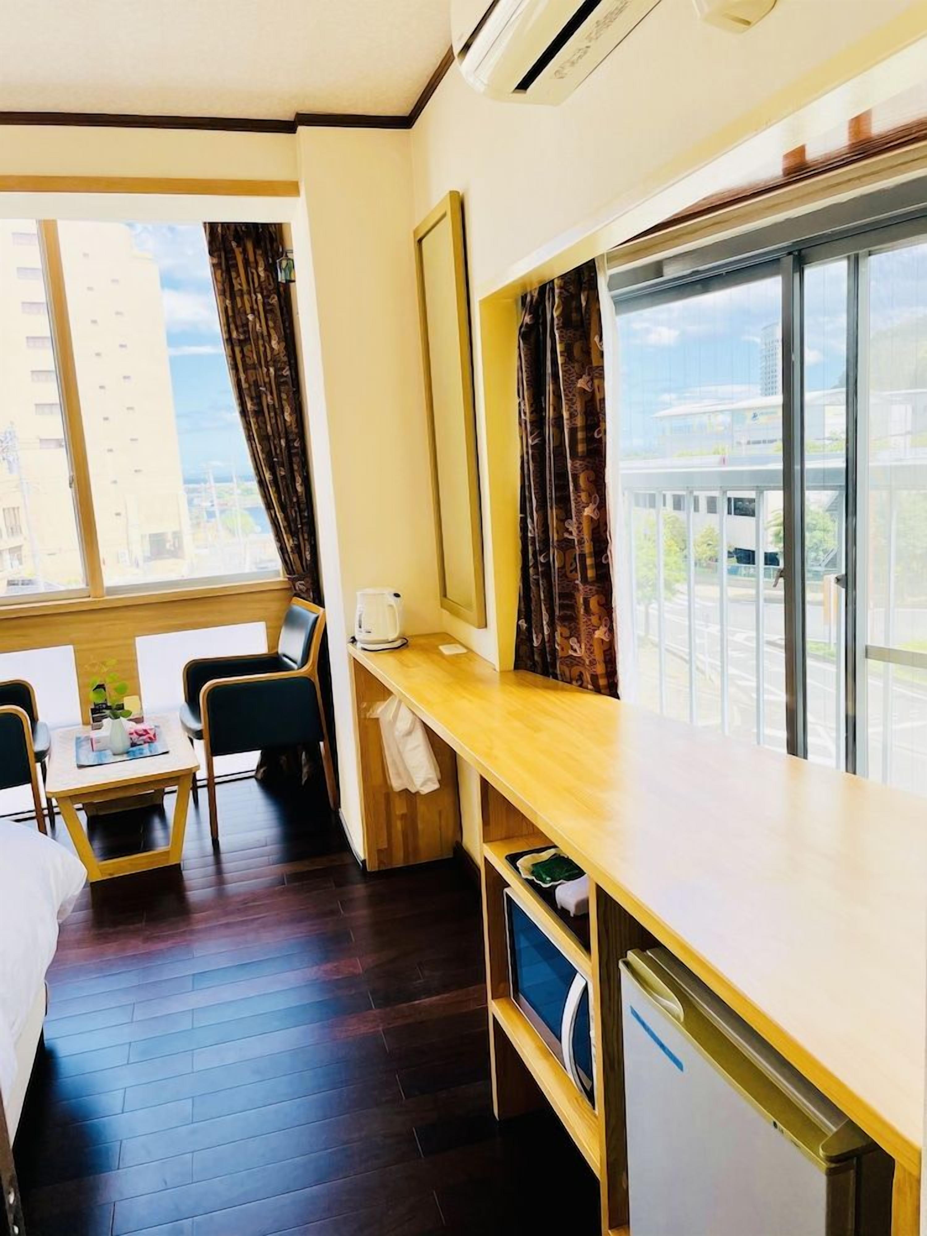 Habitación de hotel con cama, escritorio, silla, ventana con cortinas, una pequeña mesa con una planta y vistas a edificios.