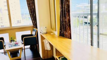 Habitación de hotel con cama, escritorio, silla, ventana con cortinas, una pequeña mesa con una planta y vistas a edificios.