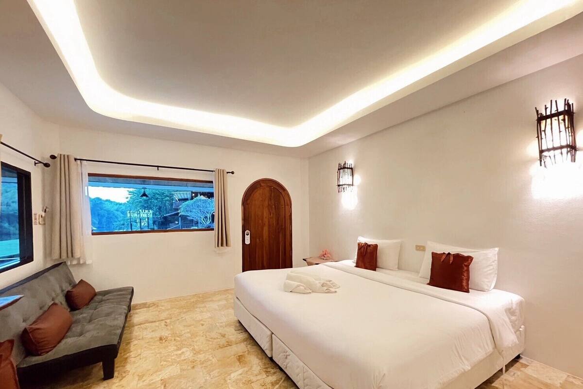 A bedroom with a large bed, a sofa, a window with a view, and a wooden door.
