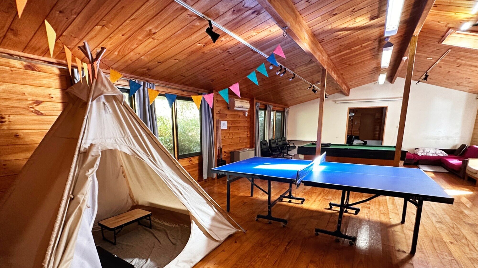 Ett tält, ett bordtennisbord och ett biljardbord i ett rum med träpanel och stora fönster.