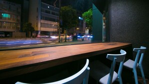 Ein Restaurant mit Holztisch und weißen Stühlen, Blick auf eine Straße mit Autos und Gebäuden im Hintergrund.