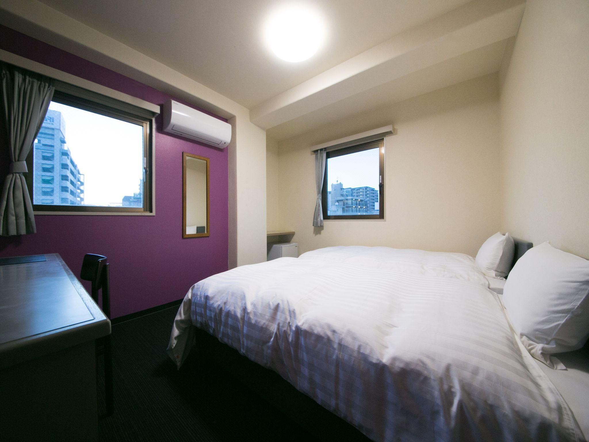 A hotel room with a bed, a desk, a chair, a window, and an air conditioning unit.
