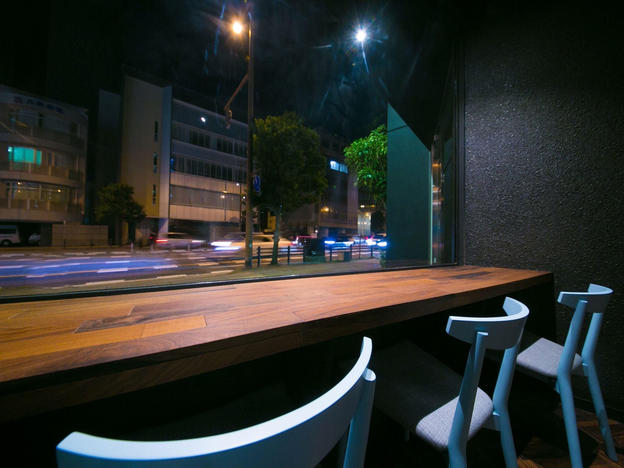 A restaurant with a wooden table and white chairs, a view of a street with cars, and buildings in the background.