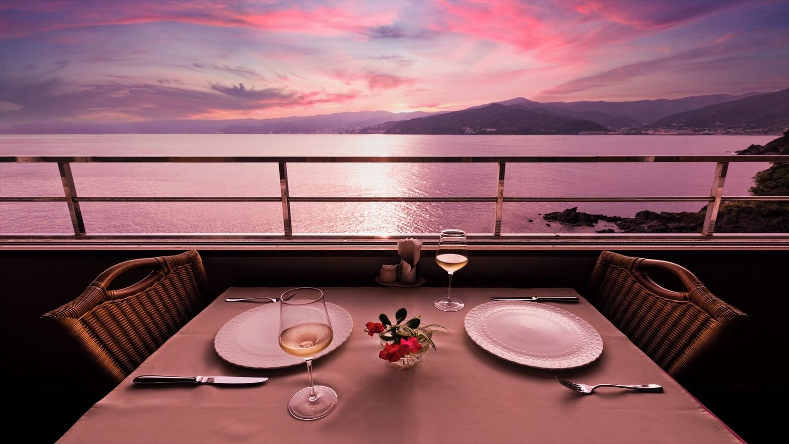 A table set for two with a view of a sunset over a body of water and mountains.