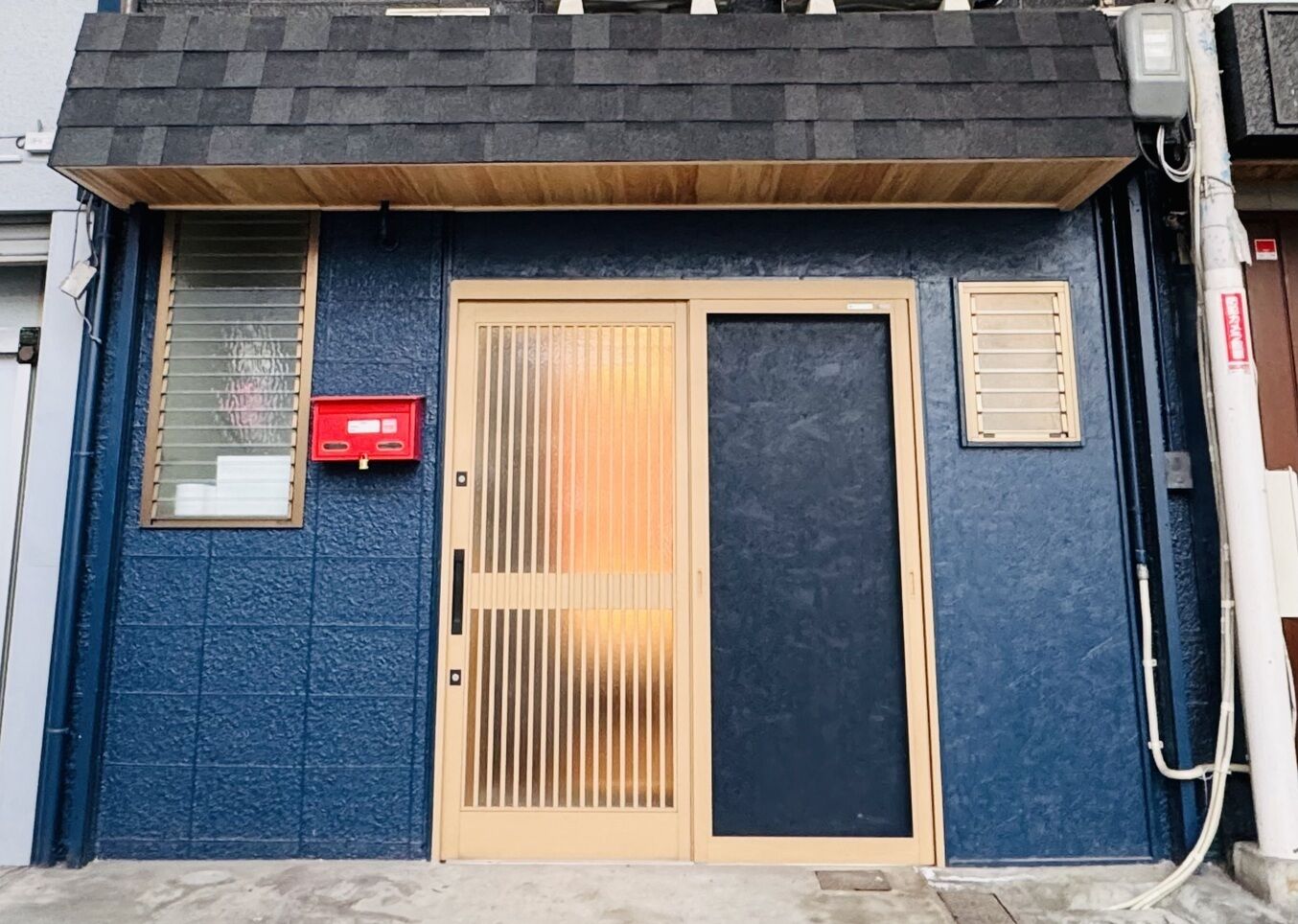 Un edificio con esterno blu e testurizzato, una porta beige con listelli verticali, una piccola finestra con persiane orizzontali e una cassetta delle lettere rossa.