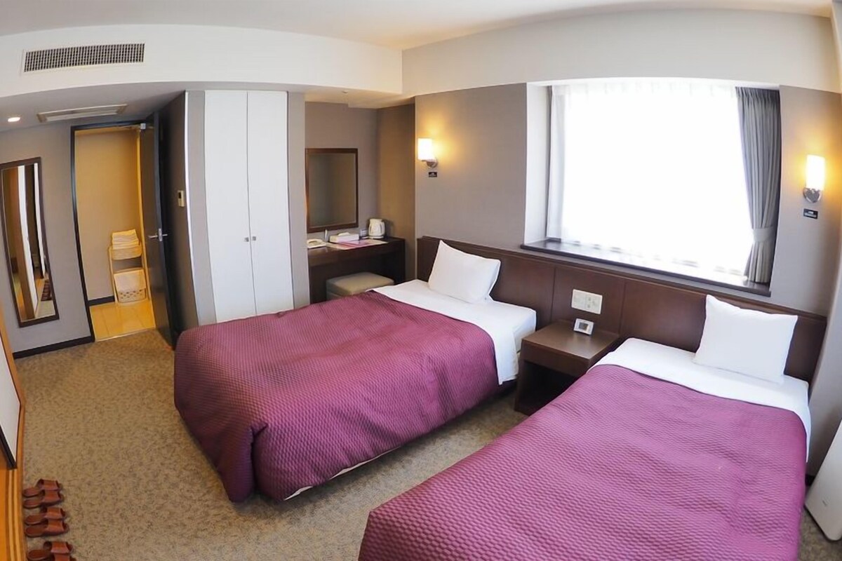 A hotel room with two beds, a wardrobe, a desk, and a window with curtains.