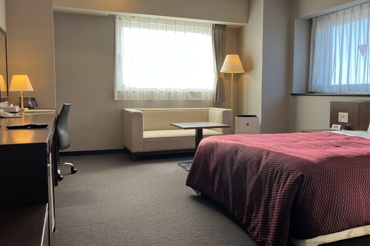 Una habitación de hotel con cama, escritorio con lámpara, silla, sofá y ventana con cortinas.