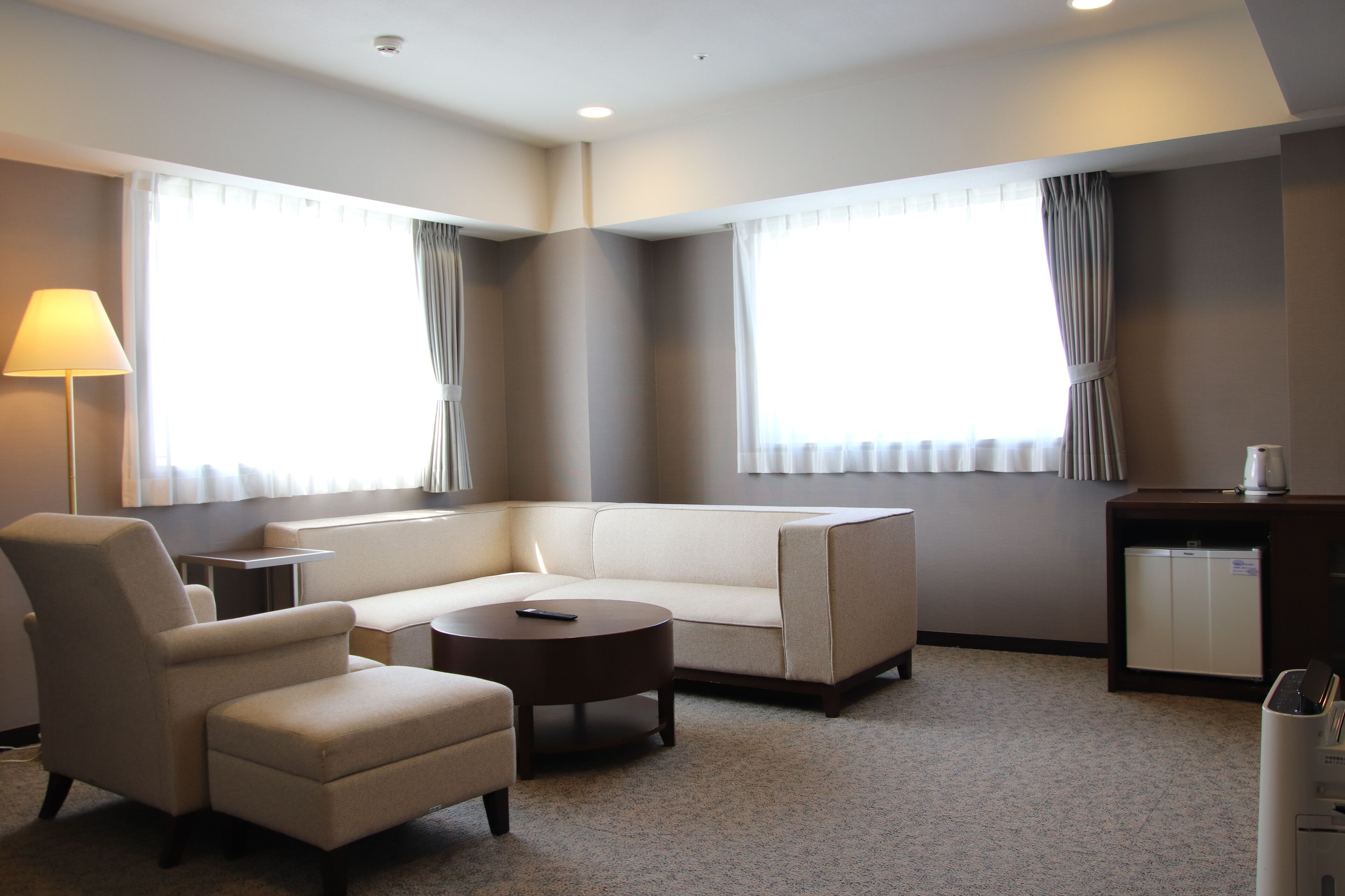 A modern hotel room with a sofa, armchair, and a small round table. There are two windows with curtains, a lamp, and a minibar.