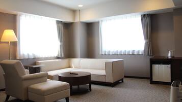 A modern hotel room with a sofa, armchair, and a small round table. There are two windows with curtains, a lamp, and a minibar.