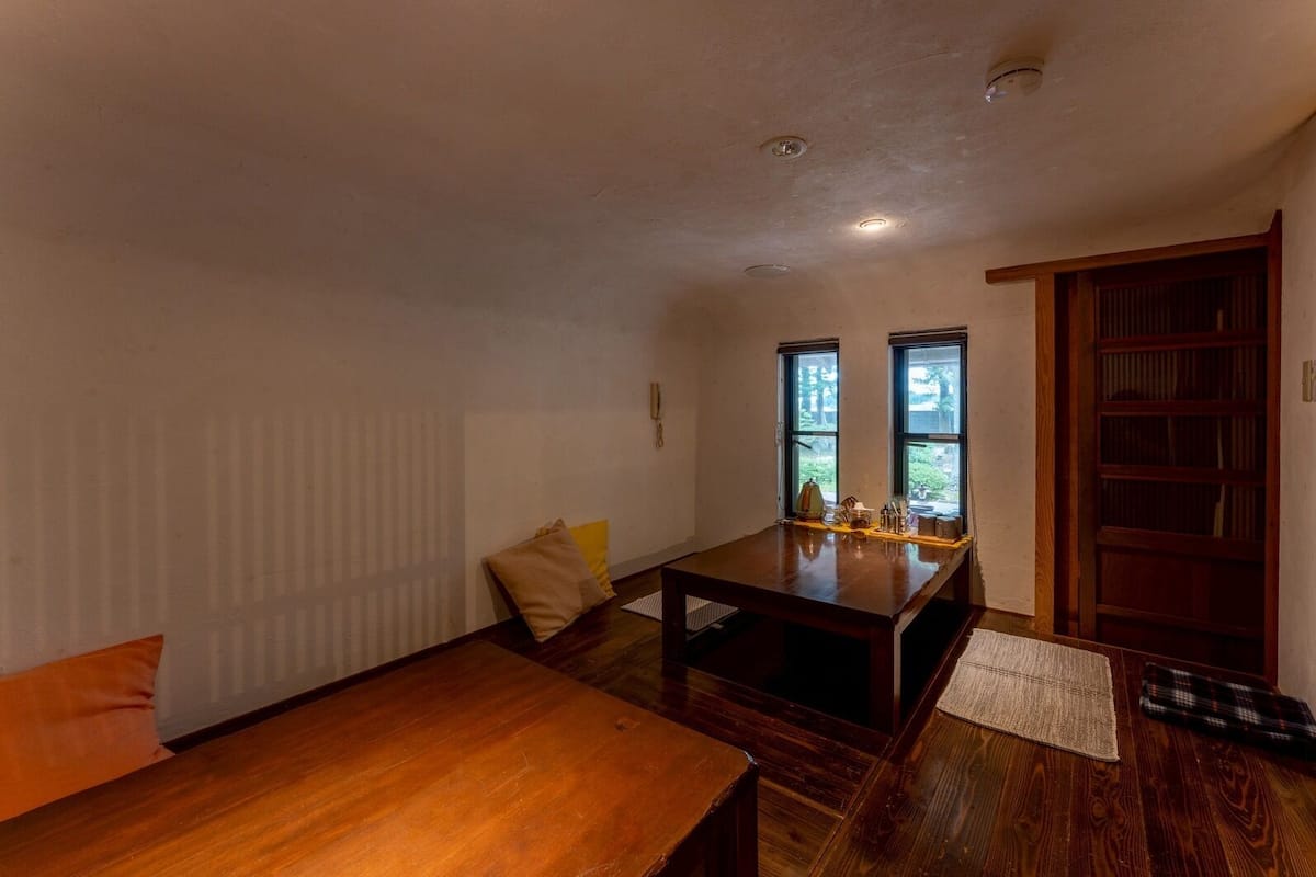 A room with a wooden table, two windows, a radiator, and a bookshelf.