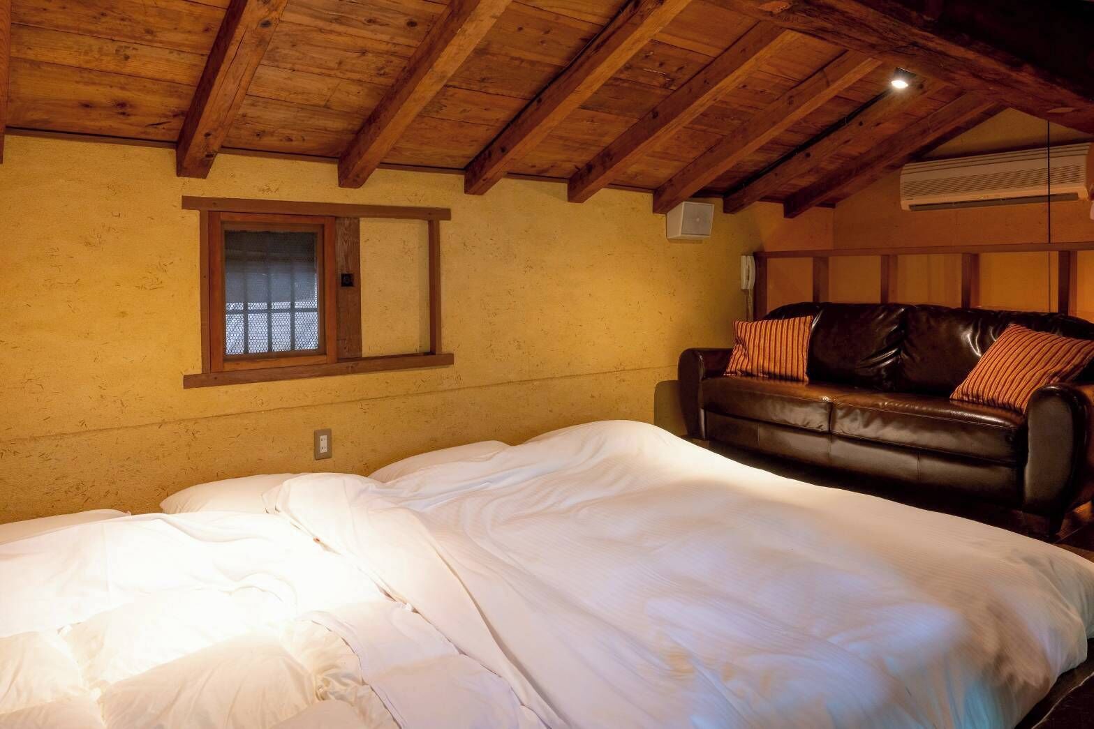 A bedroom with a wooden ceiling, a bed with white bedding, a leather sofa with pillows, and a window with a wooden frame.