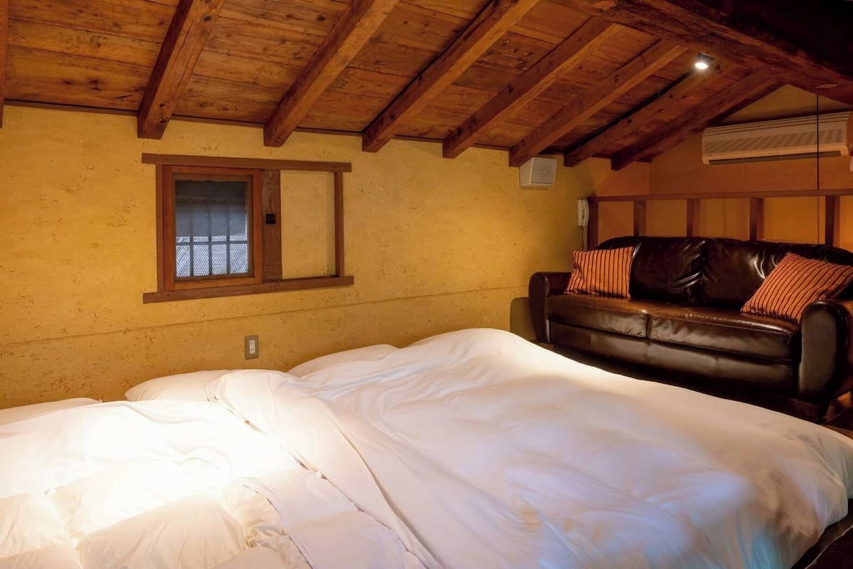 A bedroom with a wooden ceiling, a bed with white bedding, a leather sofa with pillows, and a window with a wooden frame.