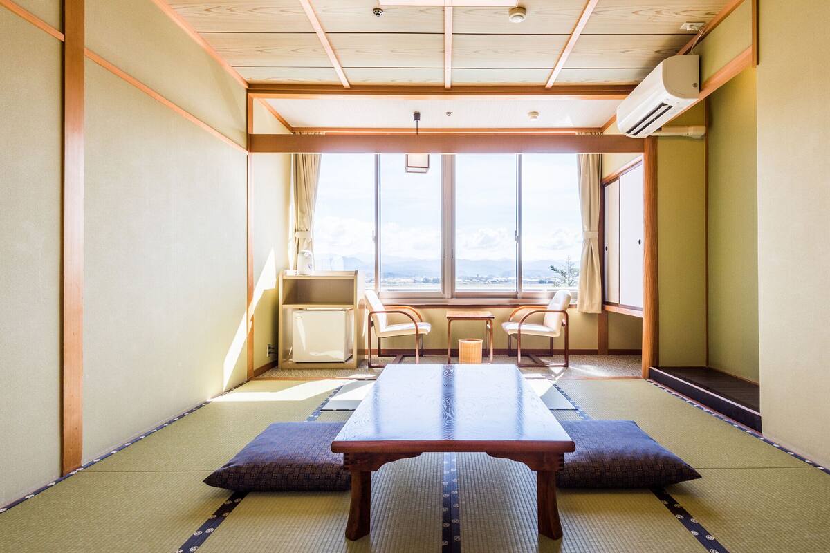 Una habitación japonesa tradicional con suelo de tatami, una mesa baja, dos sillas y vistas amplias a través de grandes ventanales.