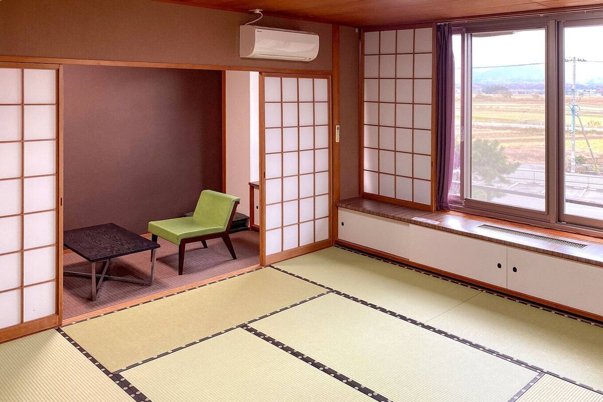 Ein Raum mit Tatami-Matten, einer Schiebetür, einem grünen Stuhl, einem kleinen Tisch und einem Fenster mit Blick ins Grüne.