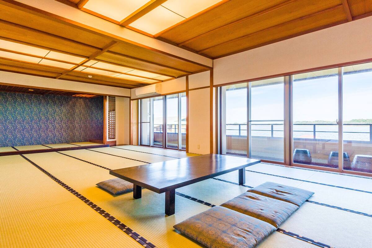 Una habitación con esteras de tatami, una mesa baja y cojines, ventanas amplias con vistas a un cuerpo de agua y un techo de madera.