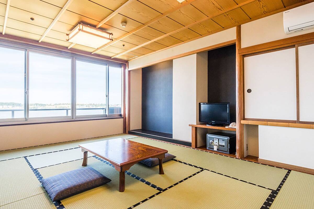 Ein traditioneller japanischer Raum mit Tatami-Bodenbelag, einem niedrigen Tisch, einem Fernseher und Blick durch große Fenster.