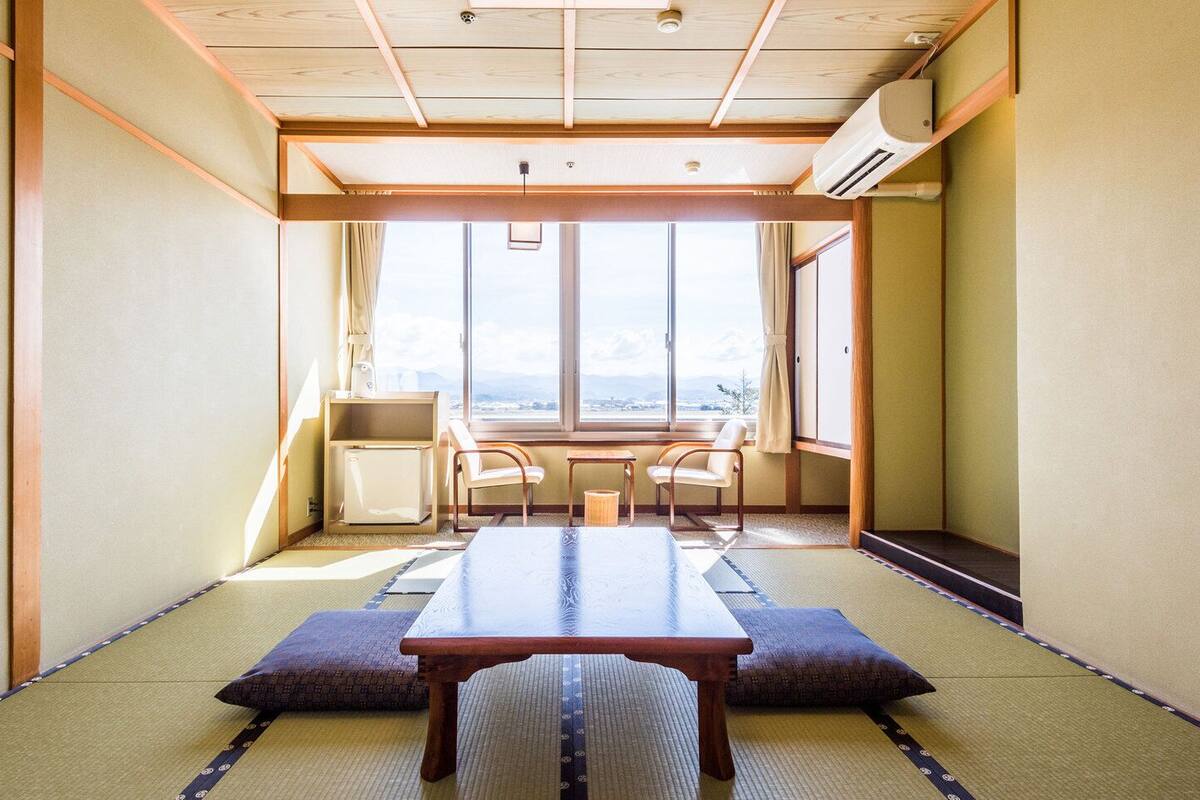 Ein traditioneller japanischer Raum mit Tatami-Boden, einem niedrigen Tisch, zwei Stühlen und Blick durch große Fenster.