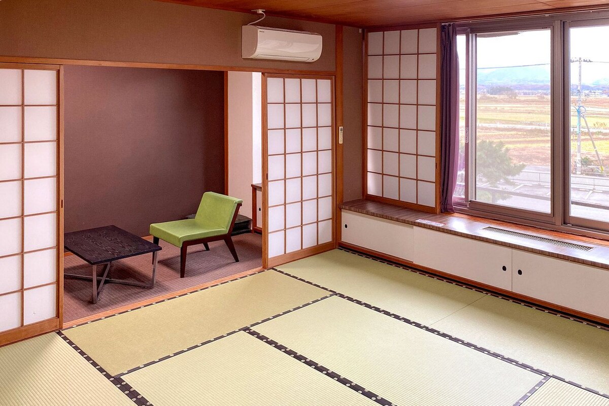 A tatami mat room with a sliding door, a green chair, a small table, and a window with a view of the countryside.