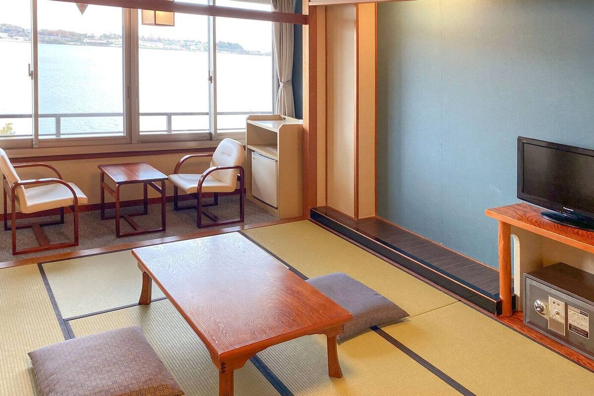 Una habitación con suelo de tatami, una mesa de madera, dos sillas, un televisor y vista a un cuerpo de agua.