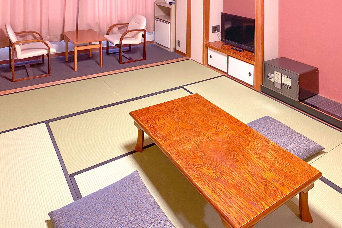 Una habitación con esteras de tatami, una mesa de madera, dos sillas, un televisor y un refrigerador pequeño.