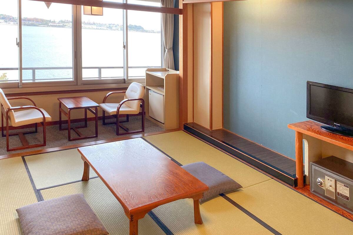 Una habitación con esteras de tatami, una mesa de madera, dos sillas, un televisor y vista a un cuerpo de agua.