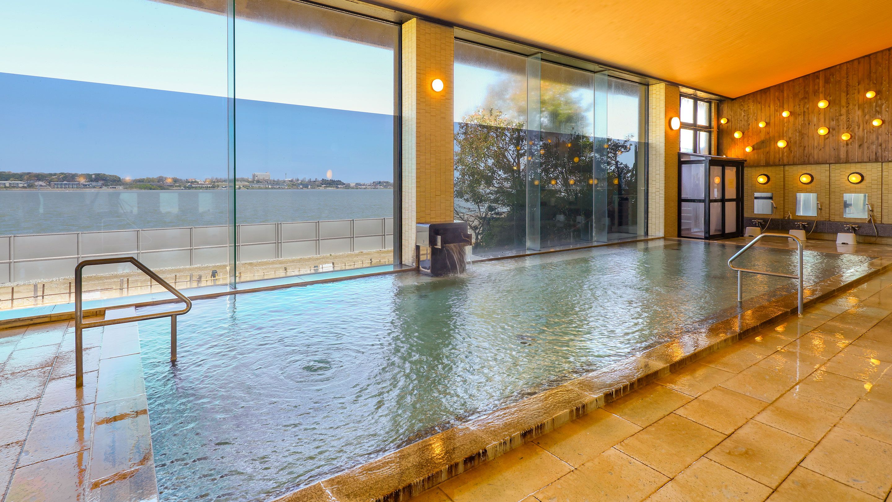 An indoor swimming pool with a view of the water body outside, a glass partition, and a wooden wall with lights.