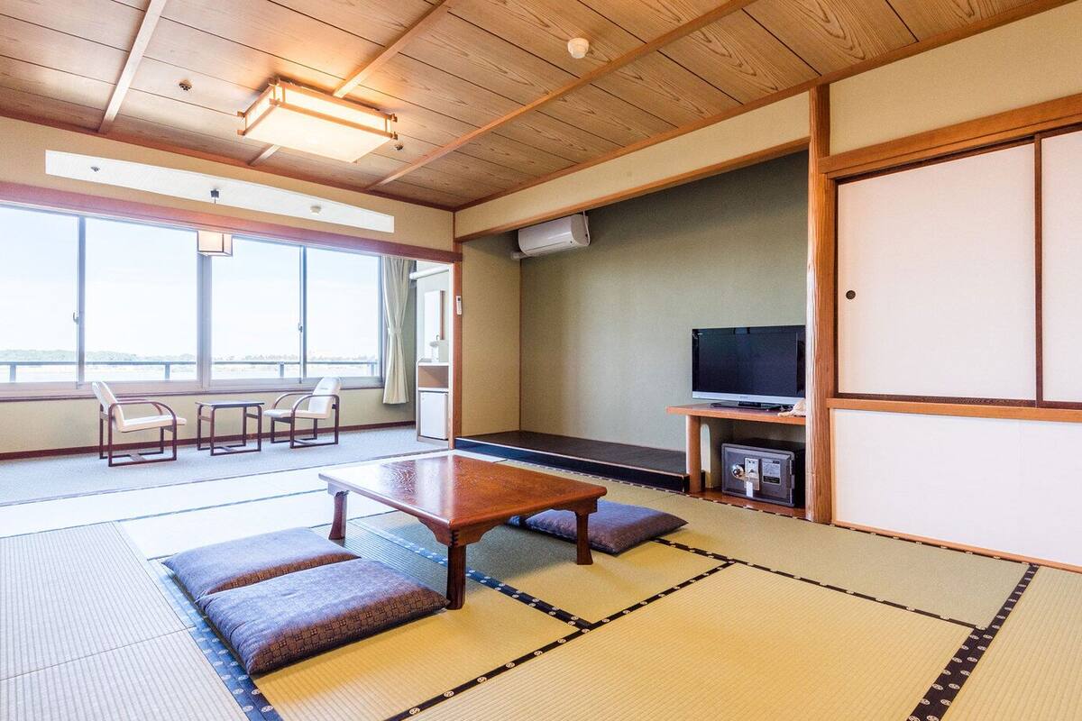 Ein traditioneller japanischer Raum mit Tatami-Bodenbelag, einem niedrigen Tisch, Kissen, einem Fernseher und Schiebetüren.