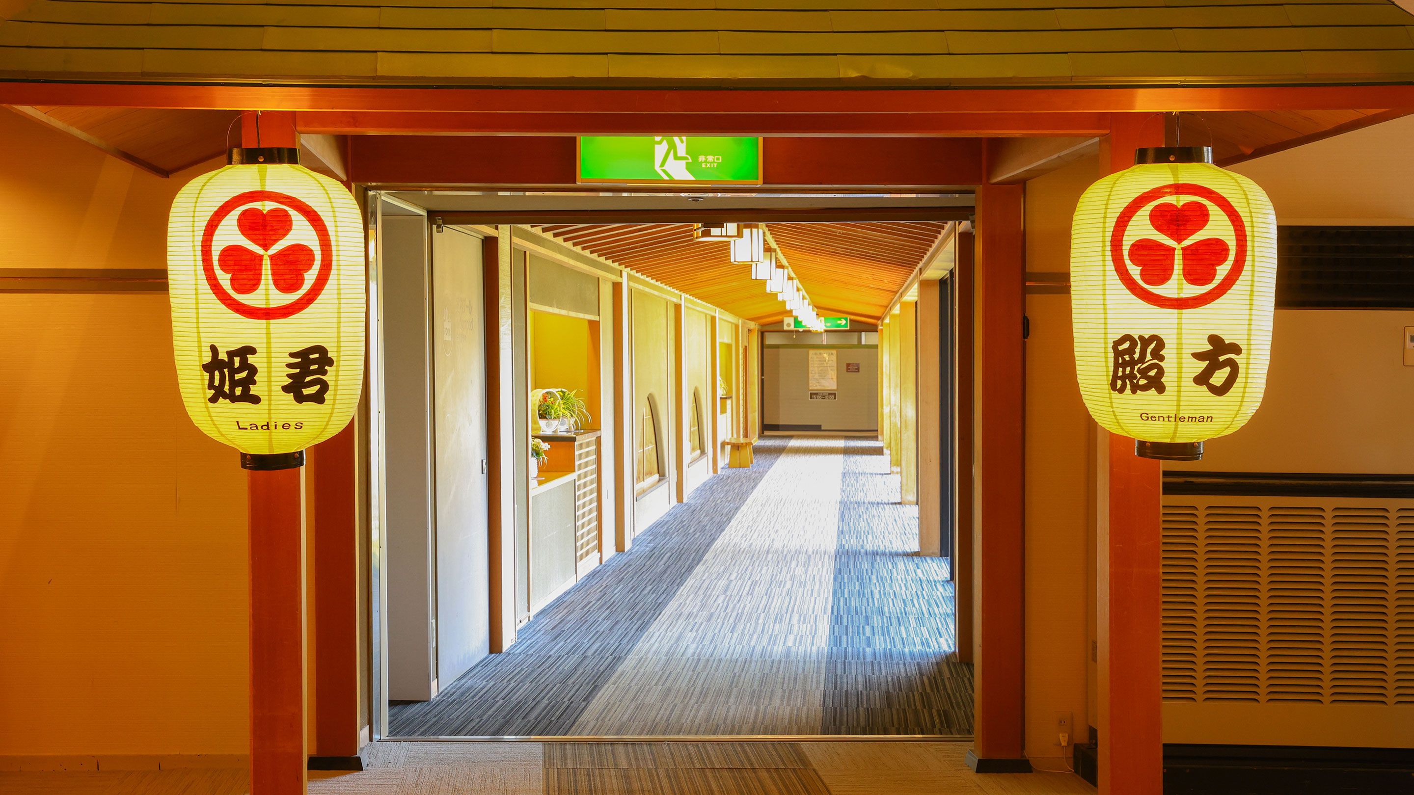 A hotel corridor with traditional Japanese lanterns, a clear exit sign, and a carpeted floor.