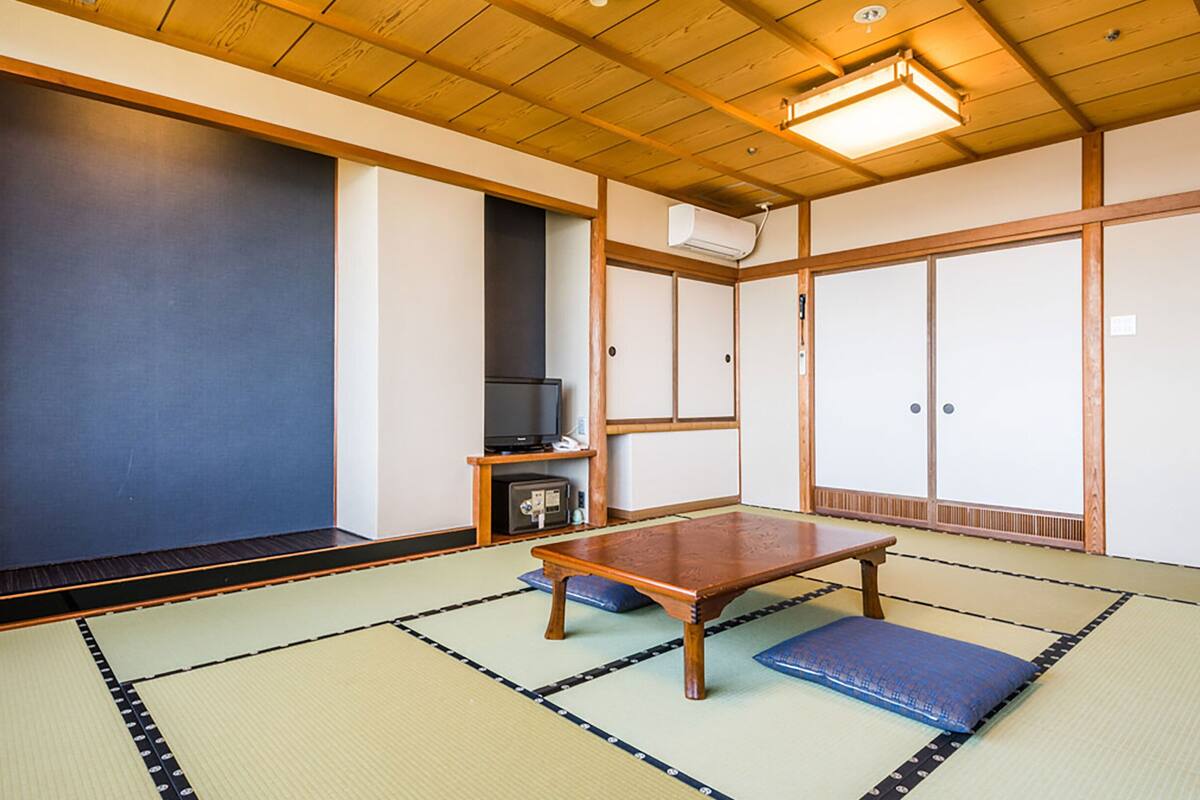 Ein Raum mit Tatami-Matten, einem niedrigen Holztisch, einem Fernseher und einer traditionellen japanischen Schiebetür.