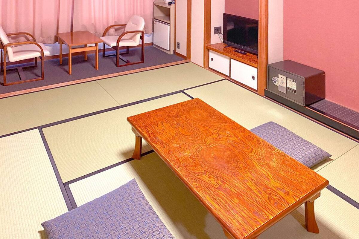 Ein Raum mit Tatami-Matten, einem Holztisch, zwei Stühlen, einem Fernseher und einem kleinen Kühlschrank.