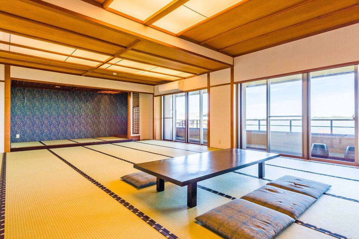Ein Raum mit Tatami-Matten, einem niedrigen Tisch und Kissen, große Fenster mit Blick auf das Wasser.