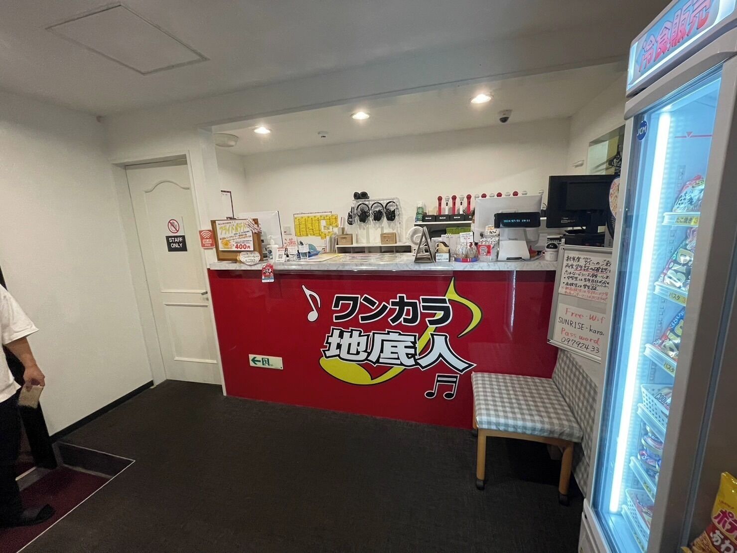 Zona de recepción con mostrador rojo, banca para sentarse, refrigerador con bebidas y un cartel que indica acceso a Wi-Fi.
