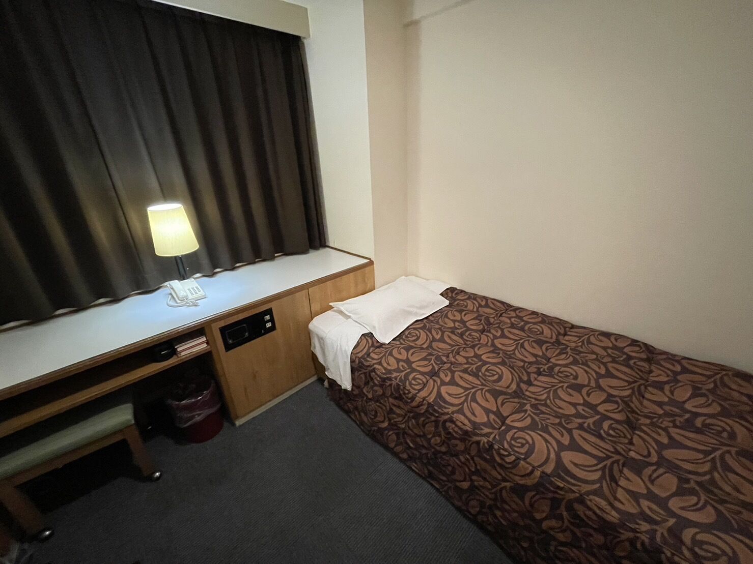 Una habitación de hotel pequeña con una cama individual, un escritorio con lámpara y una ventana con cortinas.
